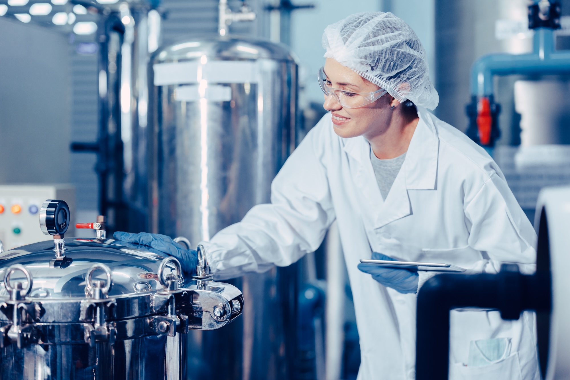 Food and beverage manufacturing professional in cleanroom attire monitoring stainless steel processing vessel with digital tablet ensuring quality control and process optimization